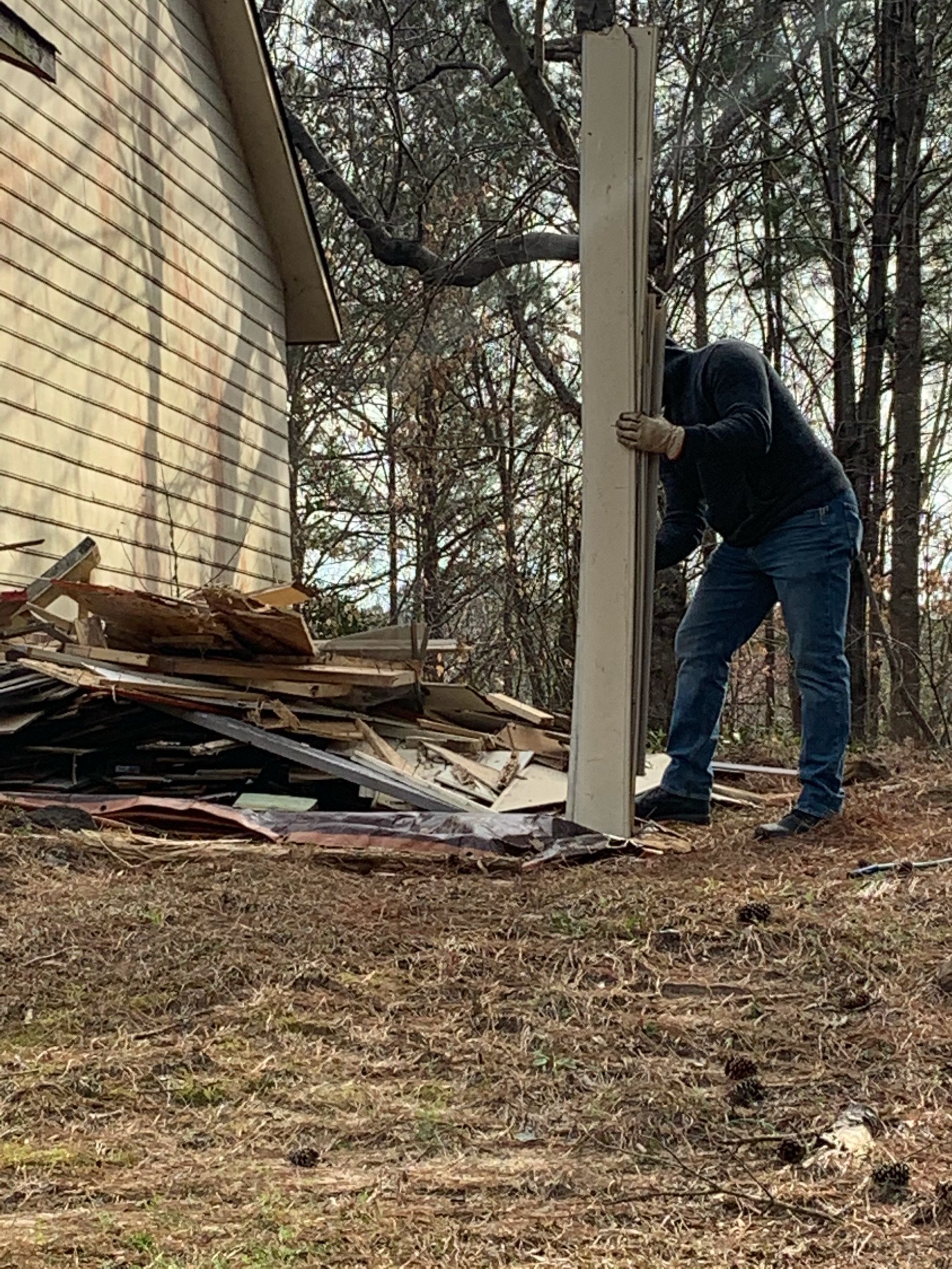 Man outside, holding a vertical beam near a building, debris on ground. Brown grass, trees in background.