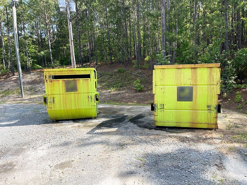 Two weathered, lime-green dumpsters sit on a gravel surface in front of a forest.