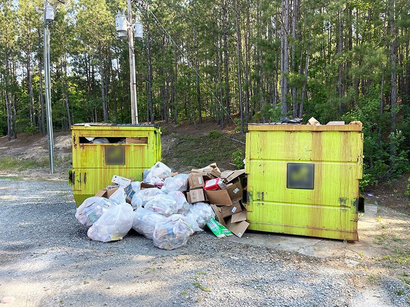 Two overflowing yellow dumpsters with trash bags and cardboard boxes on a gravel area in front of trees.