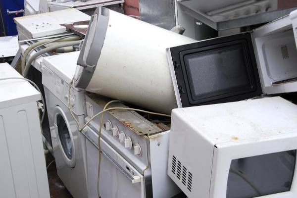 Pile of discarded white appliances: washing machine, oven, microwave, and refrigerator.