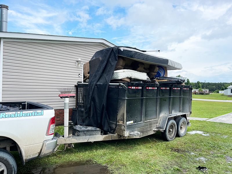 A truck towing a trailer overflowing with junk, parked next to a house on a cloudy day.