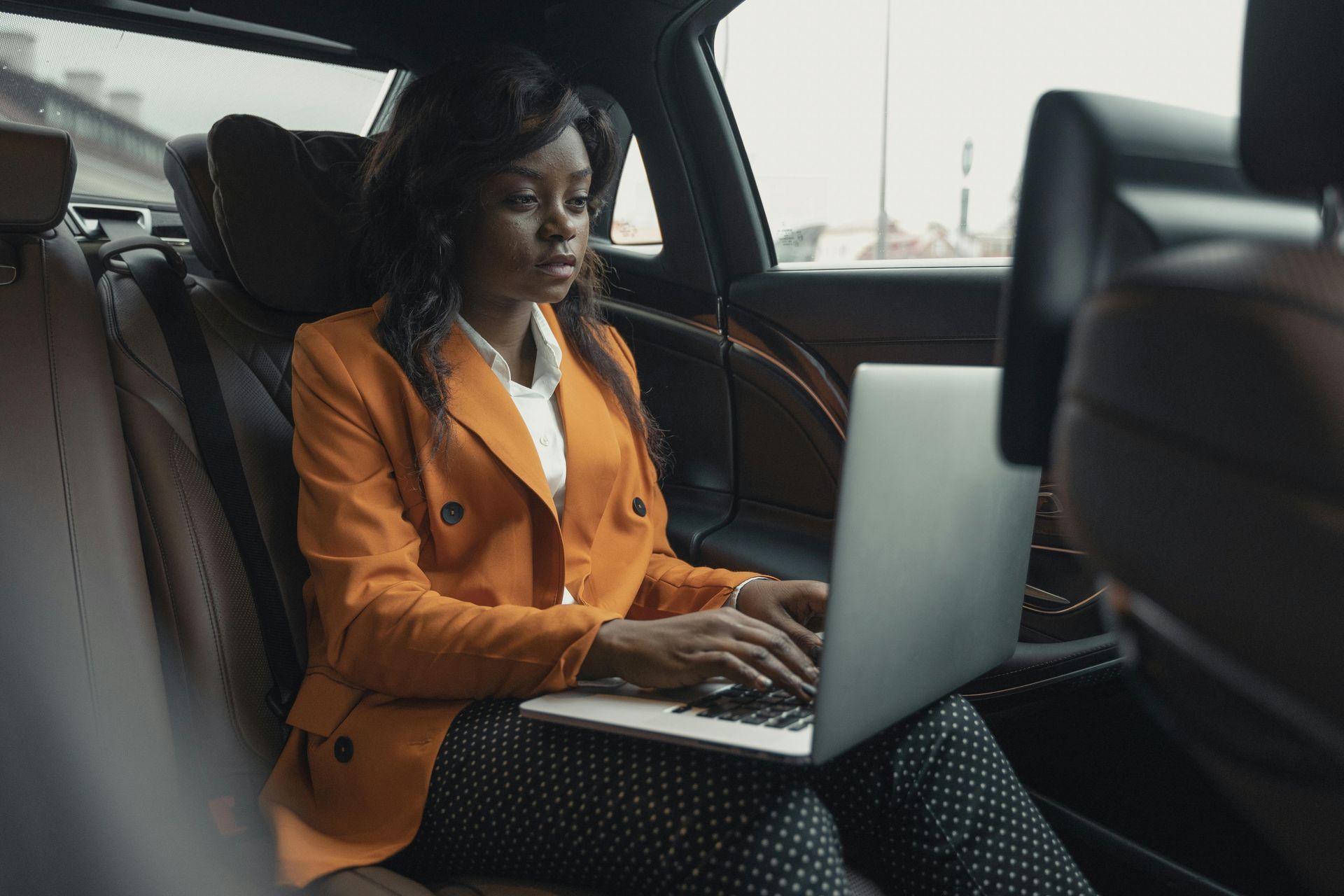 A woman is sitting in the back seat of a car using a laptop computer.