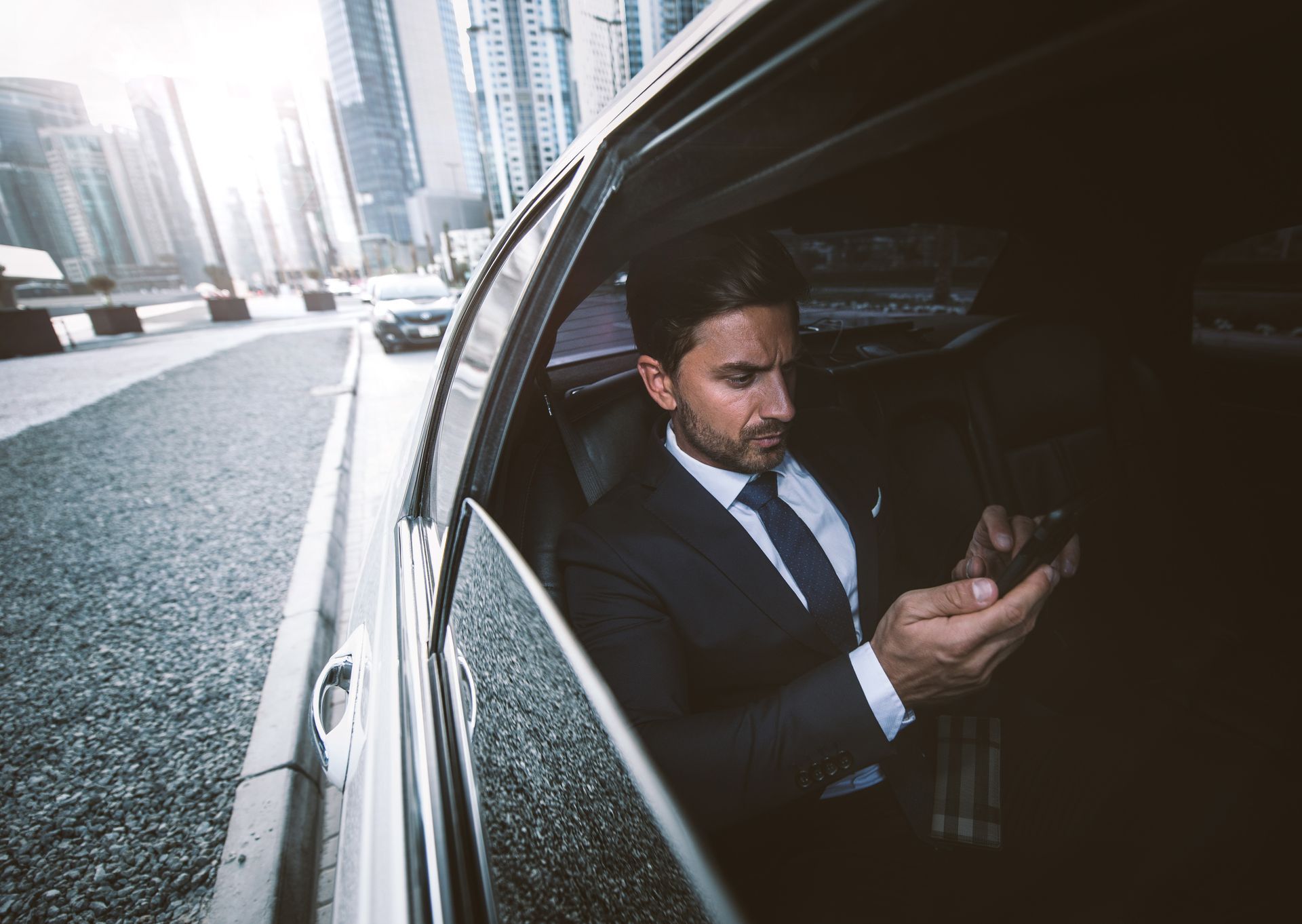 A man in a suit and tie is sitting in a car looking at his cell phone.