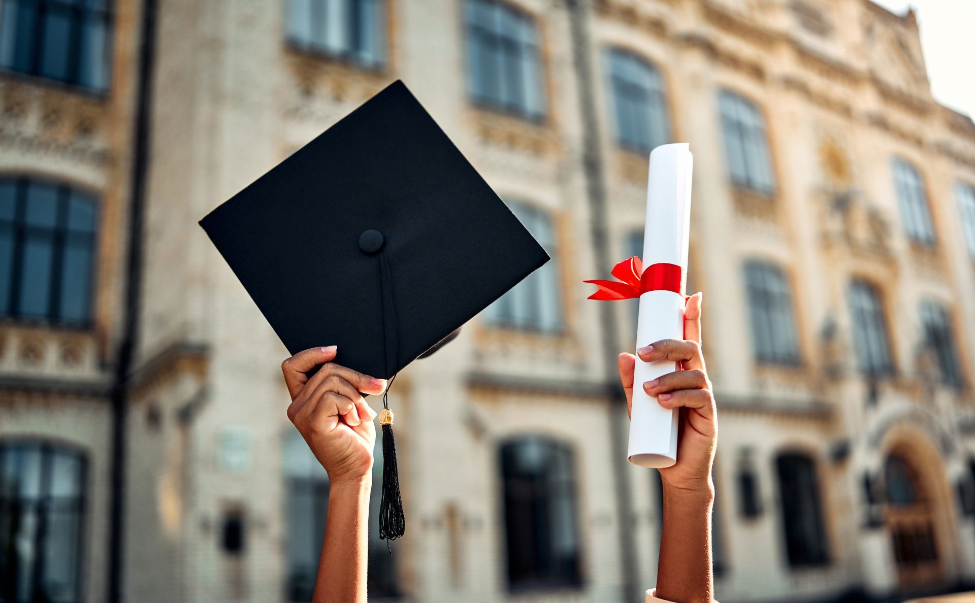 A person is holding a graduation cap and diploma in front of a building.