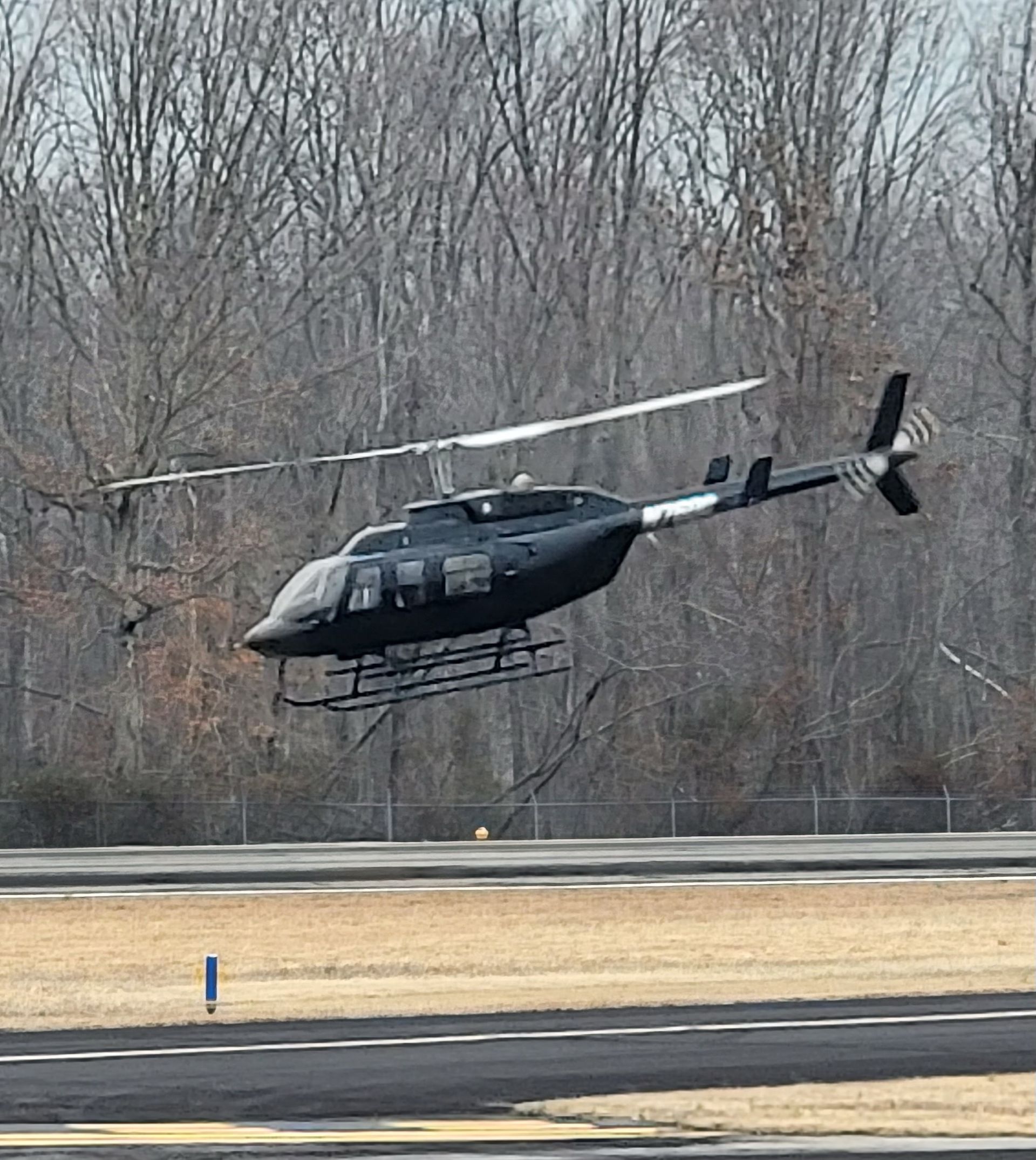 A black helicopter is flying over a runway with trees in the background