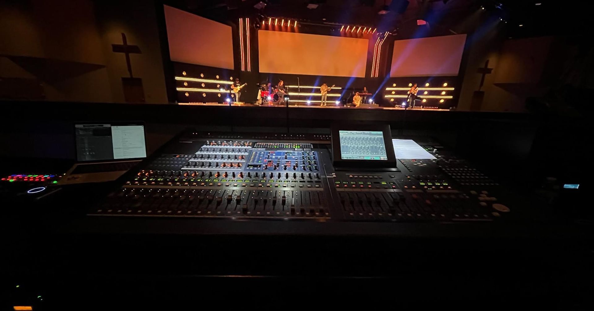 Sound mixing console in a darkened room, stage with band in the background, lit screens.