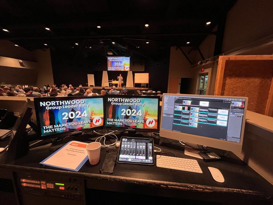 Control room setup for a conference with multiple screens displaying graphics and a live feed of the stage.