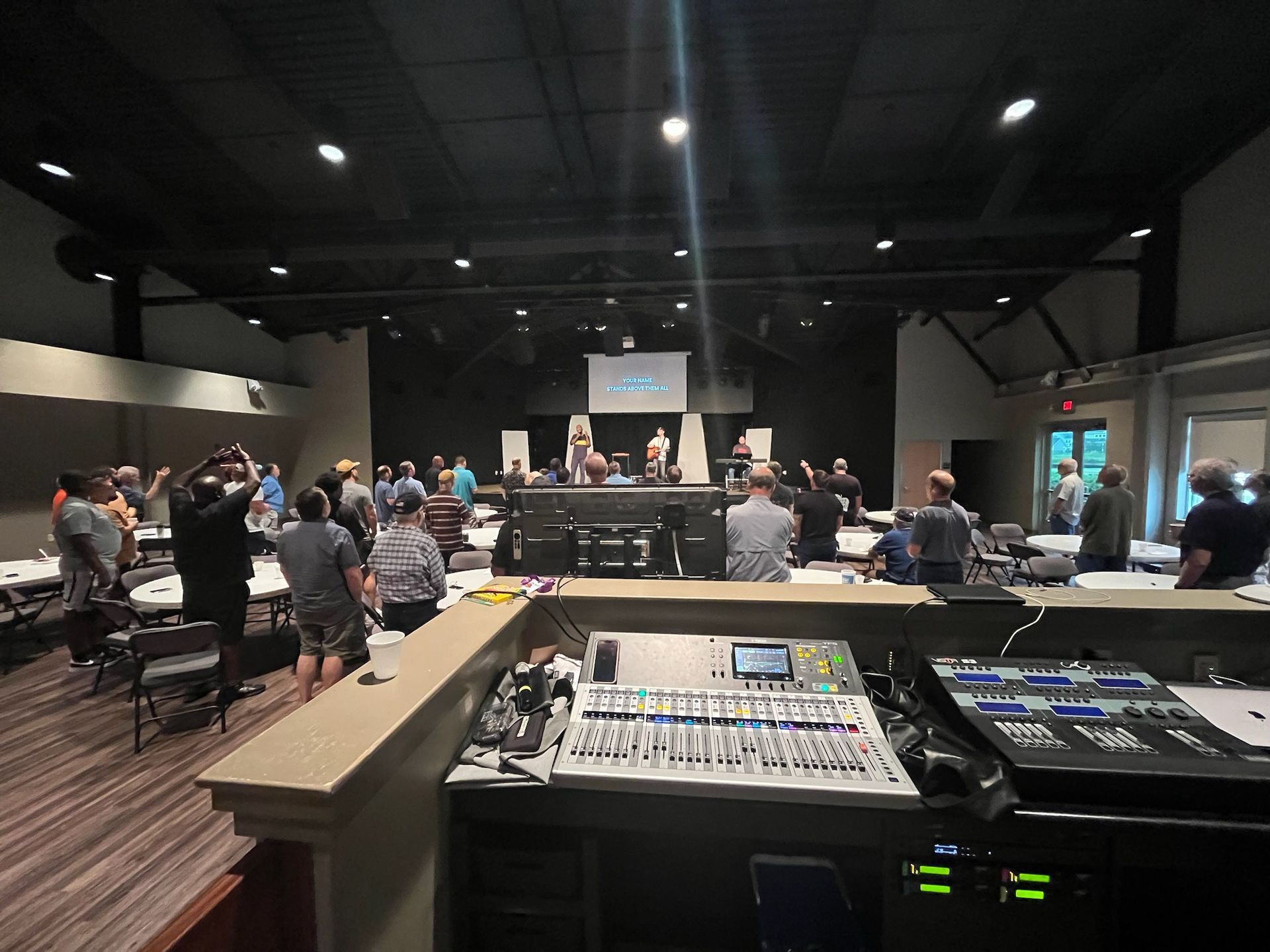 People at a meeting in a large room, with a soundboard in the foreground. Stage in the background.
