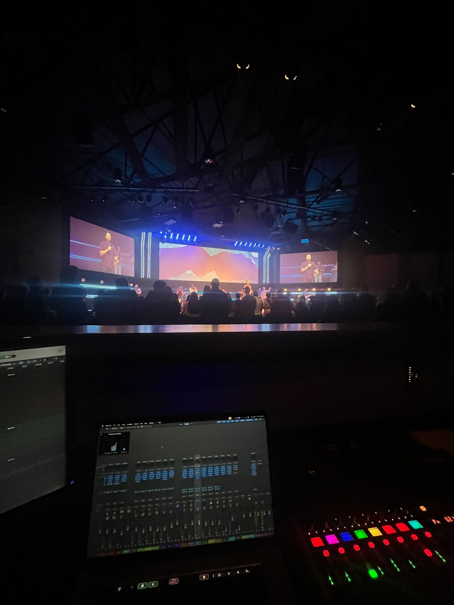 Control panel view of a stage with a screen displaying a performance. Blue, orange, and white lights illuminate the stage.