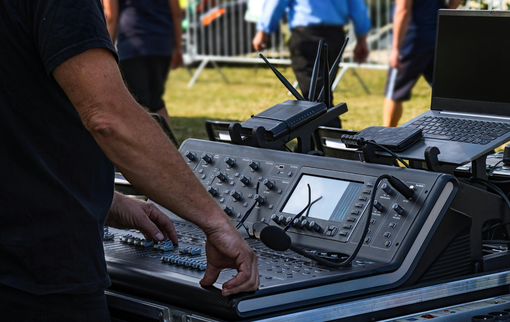 Person operating a sound mixing console outdoors; laptop and other equipment visible.