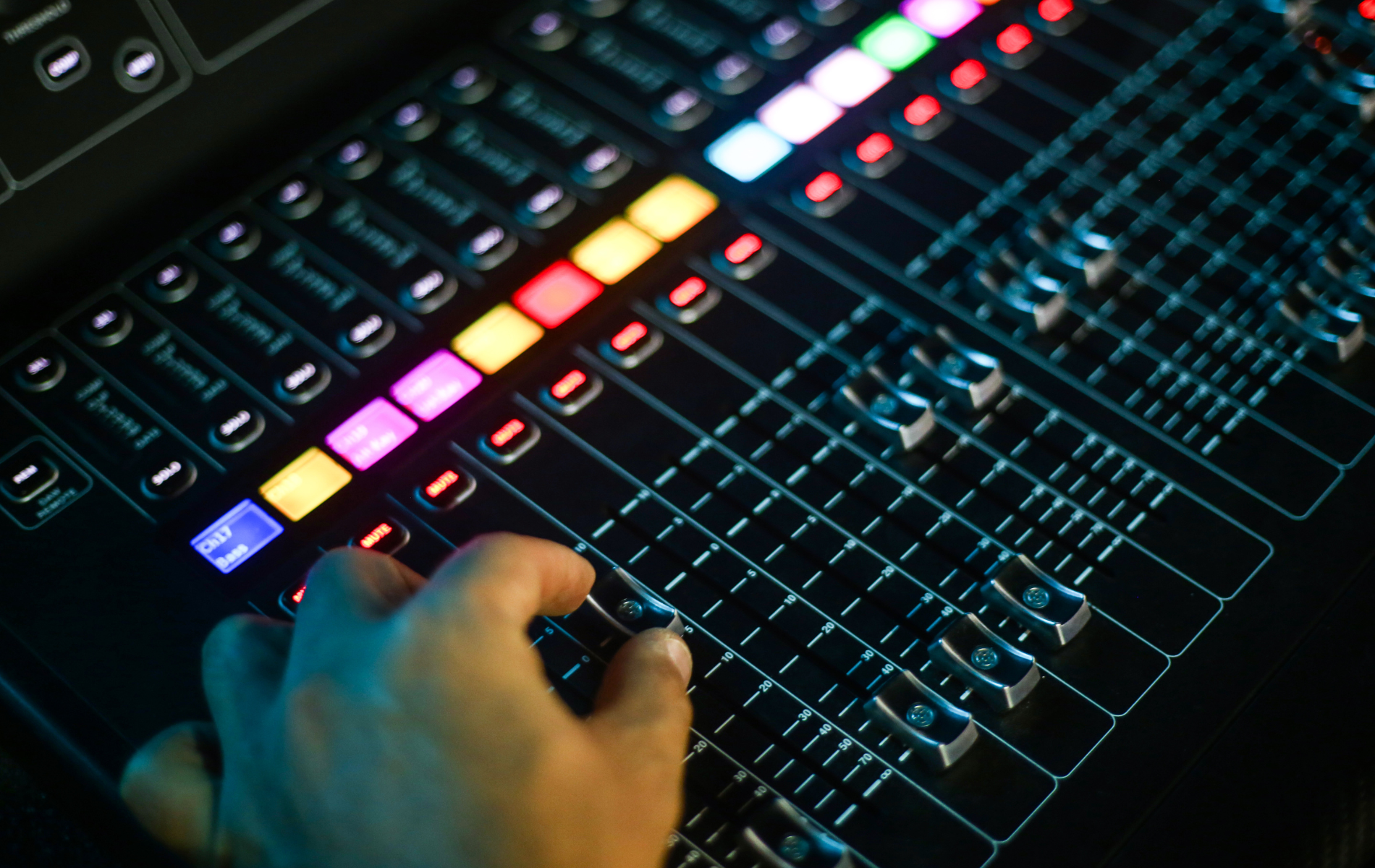 Hand adjusting fader on a black audio mixing console with illuminated controls.