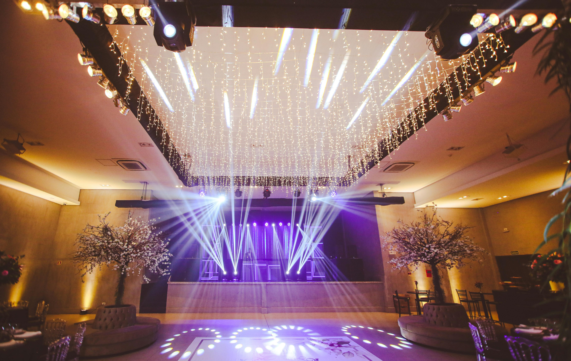 Stage with purple and white lights, decorated ceiling, and dance floor in an event hall.