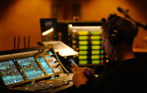 Man wearing headphones, operating audio equipment with console screens, using a phone. Dark, indoor setting.