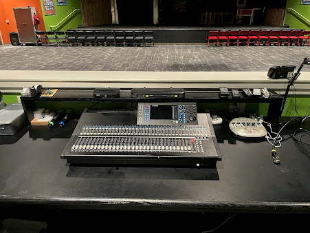 Sound mixing console on a black table in front of an empty theater stage and seating.