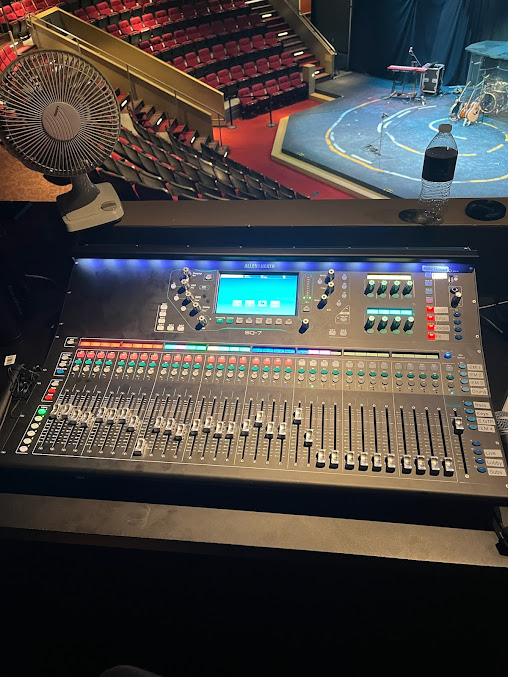 Mixing console in a theater booth overlooking the stage and audience seating.