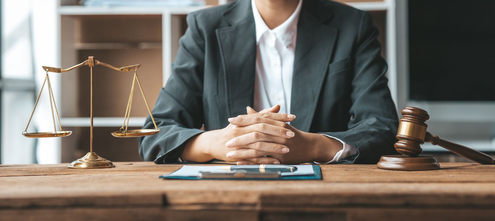 Person in a blazer sitting behind a desk with a gavel, scales of justice, and a clipboard.