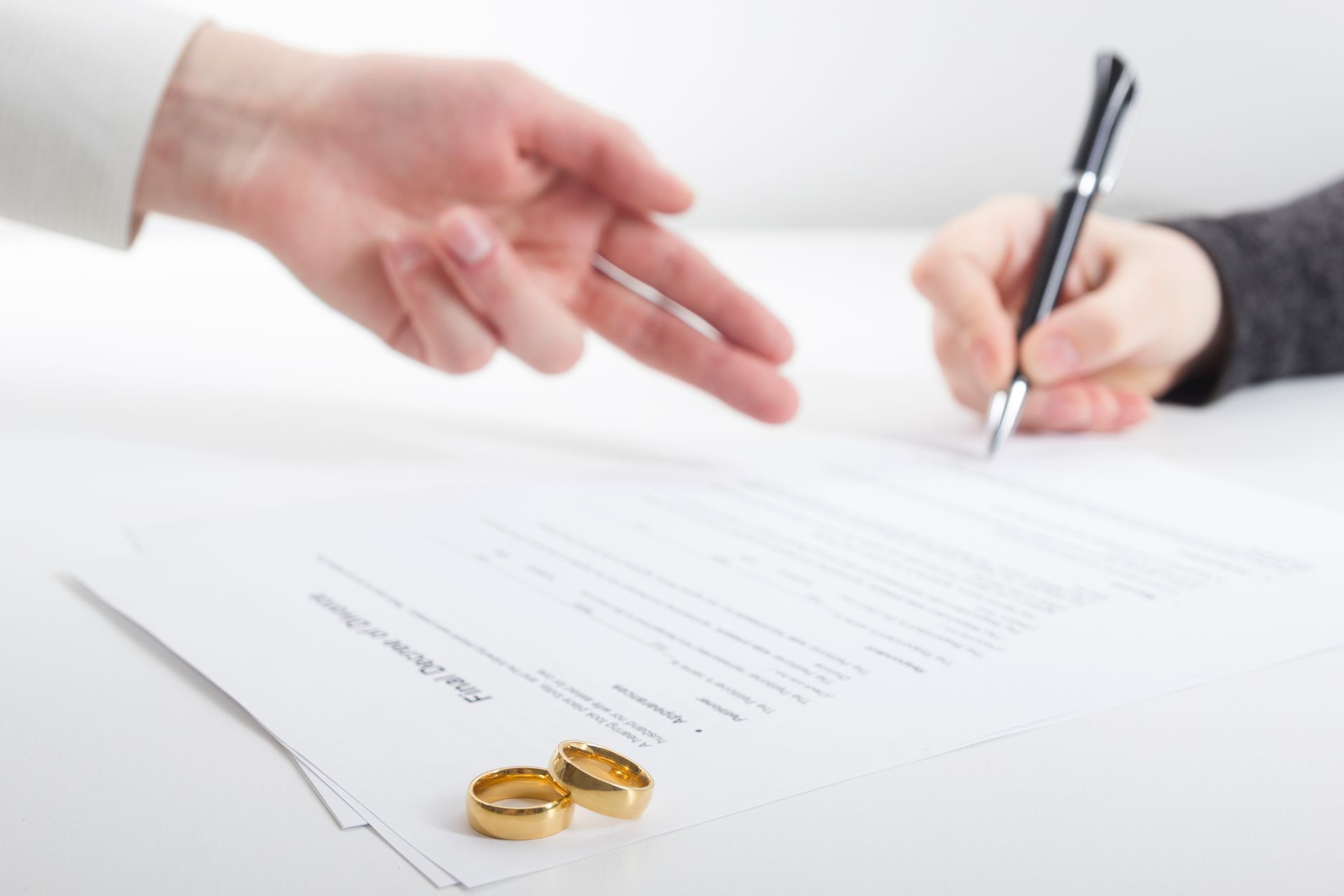 Two hands, one signing a a prenuptial agreement, another pointing. Wedding rings on the paper.