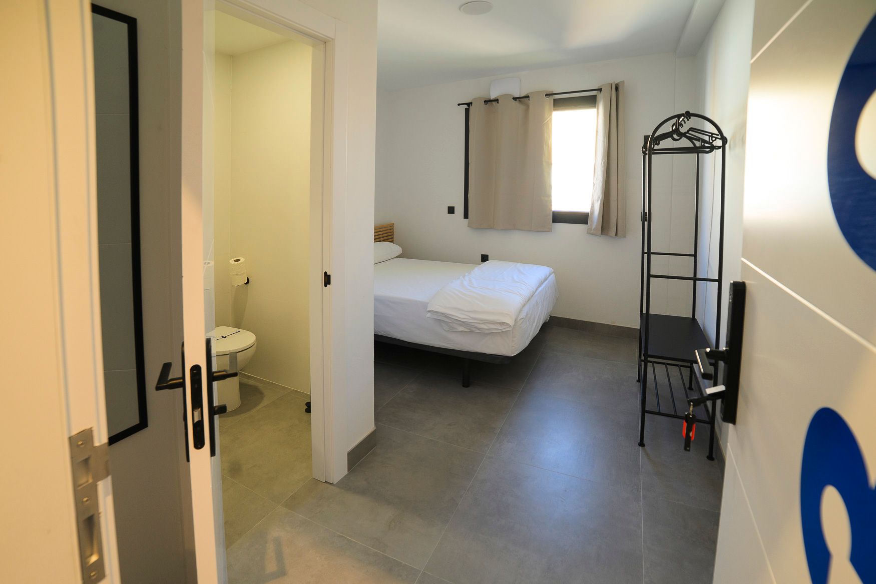 Vista de un dormitorio sencillo con baño a través de una puerta abierta. Paredes blancas, cama individual, suelo gris.