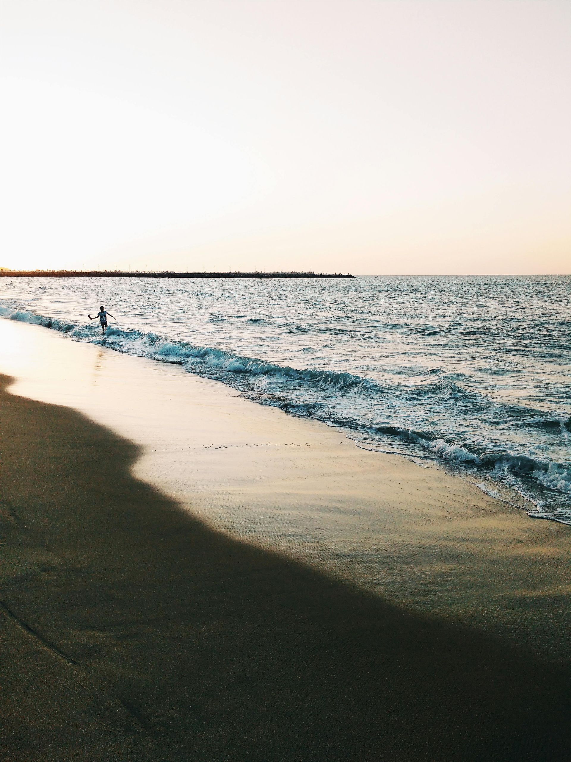 Playa al atardecer con olas rompiendo en la orilla. Una figura está de pie en el agua.