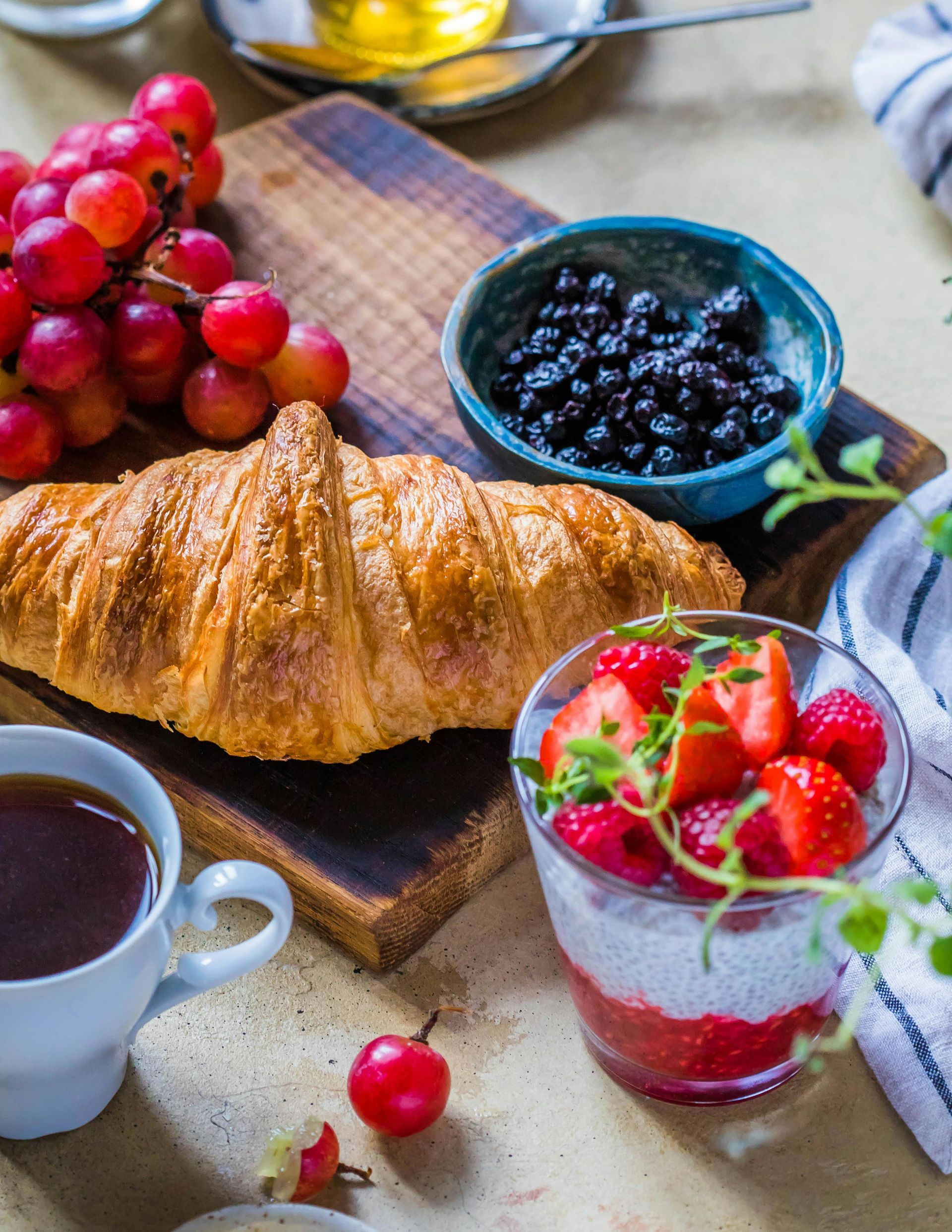 Croissant, uvas, arándanos, parfait de yogur y café sobre una tabla de madera para un desayuno luminoso.