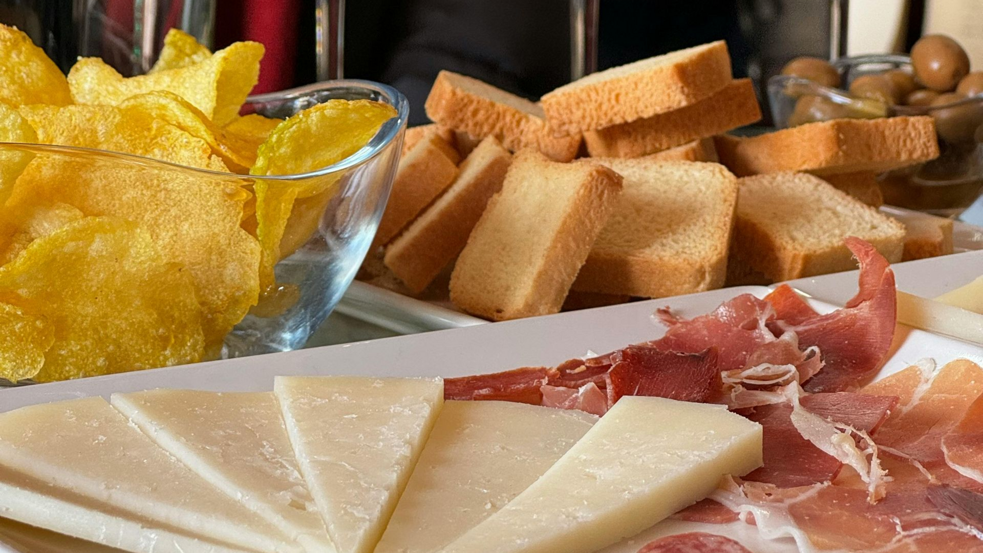 Primer plano de alimentos que incluyen queso, embutidos, galletas, patatas fritas y aceitunas.