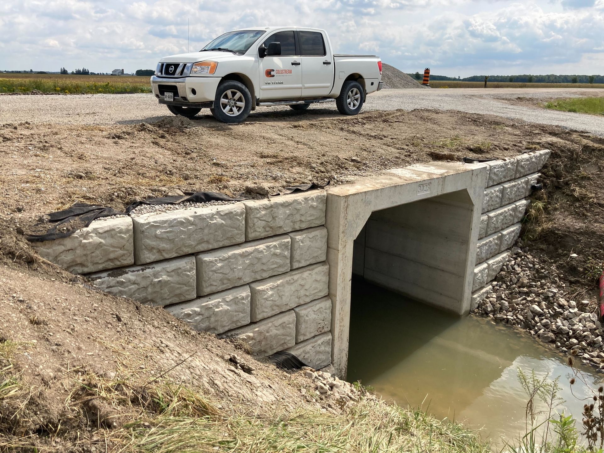 A white truck is parked on the side of a dirt road next to a concrete wall.