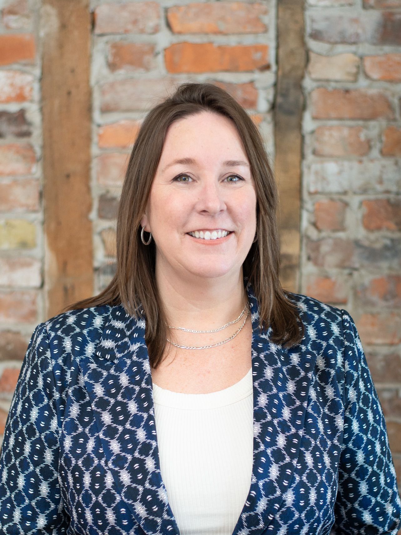 A woman in a blue jacket is smiling in front of a brick wall.