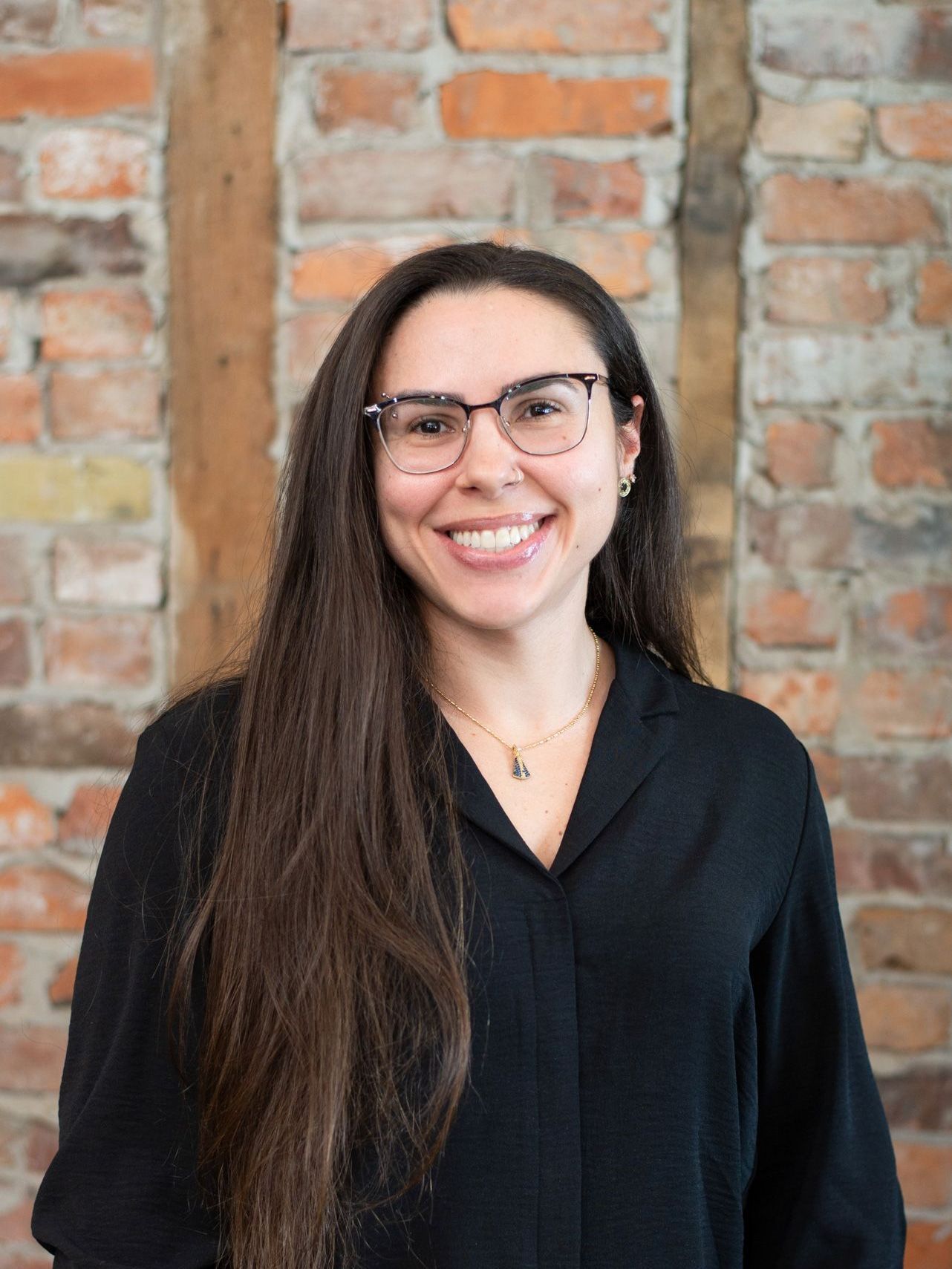 A woman wearing glasses and a black shirt is smiling in front of a brick wall.