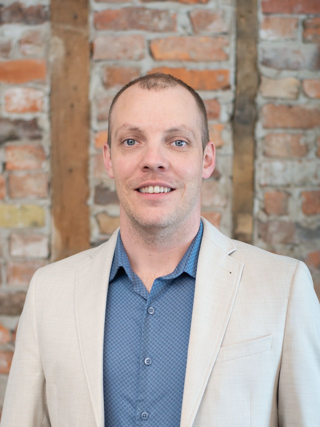 A man in a suit and blue shirt is smiling in front of a brick wall.