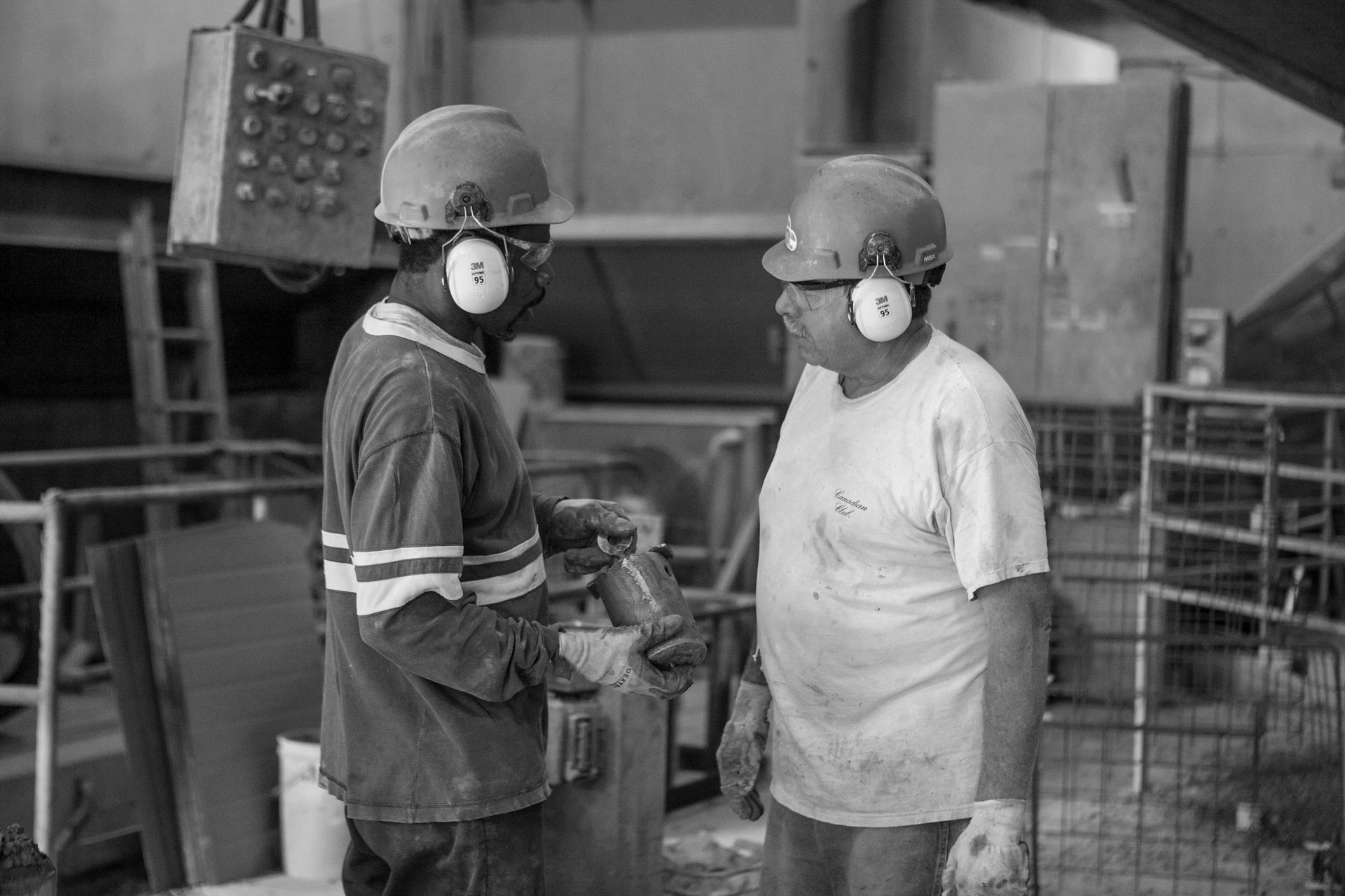 Two men wearing hard hats and ear muffs are standing next to each other in a factory.