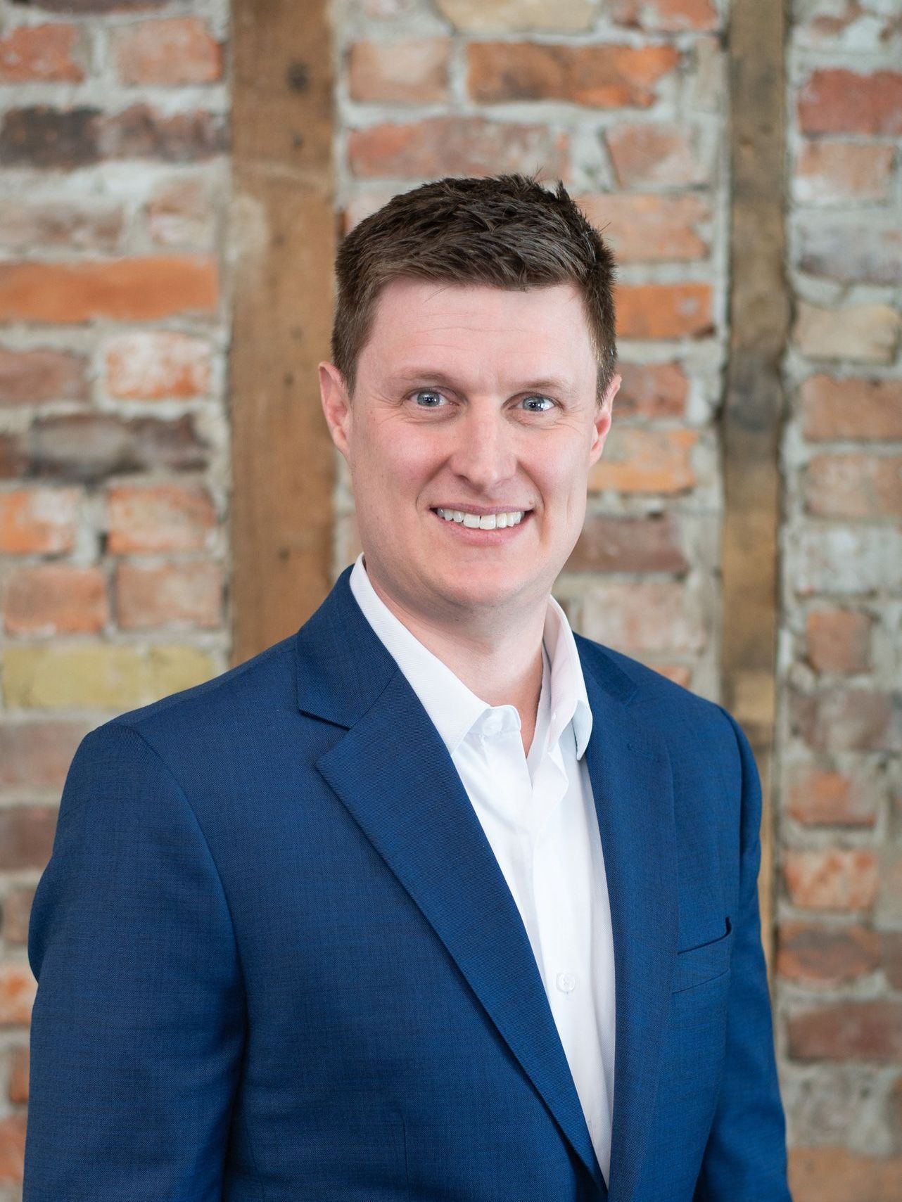 A man in a blue suit and white shirt is smiling in front of a brick wall.