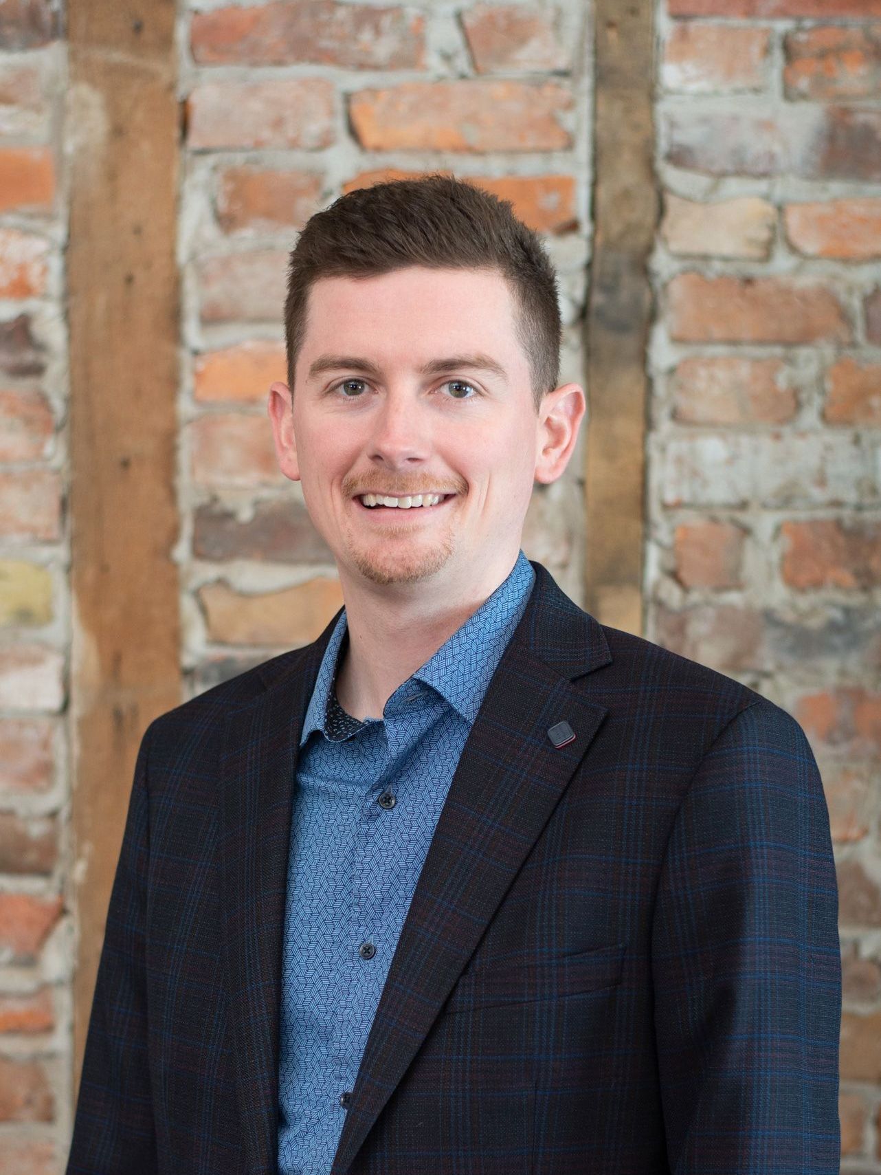 A man in a suit and blue shirt is standing in front of a brick wall.
