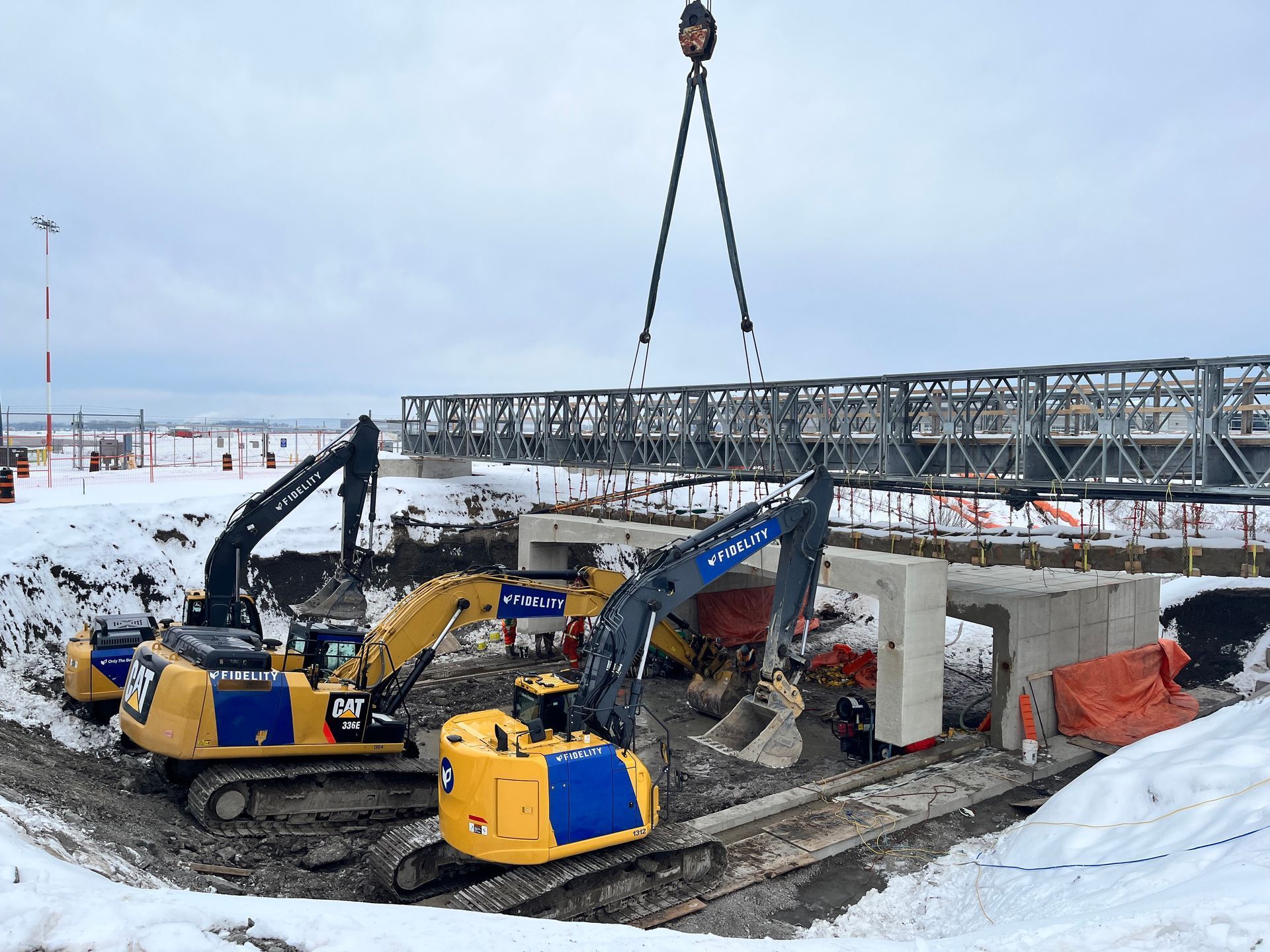 A yellow and blue excavator is lifting a bridge in the snow.
