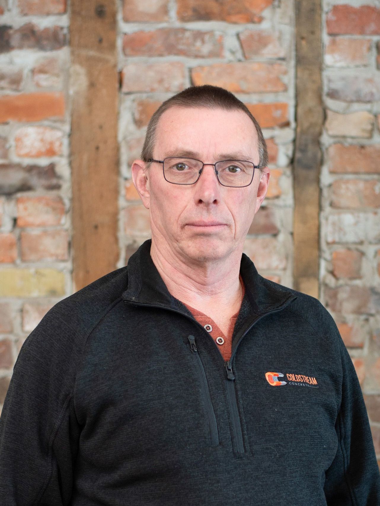 A man wearing glasses and a black sweater is standing in front of a brick wall.