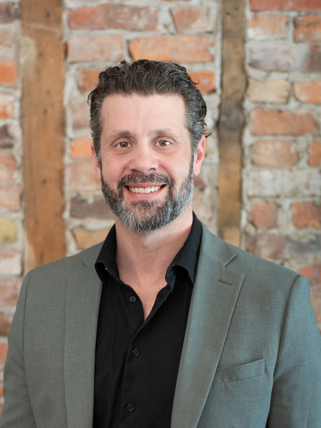 A man with a beard is wearing a suit and smiling in front of a brick wall.
