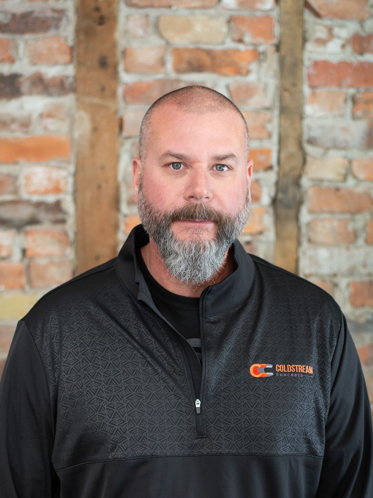 A man with a beard and a black jacket is standing in front of a brick wall.