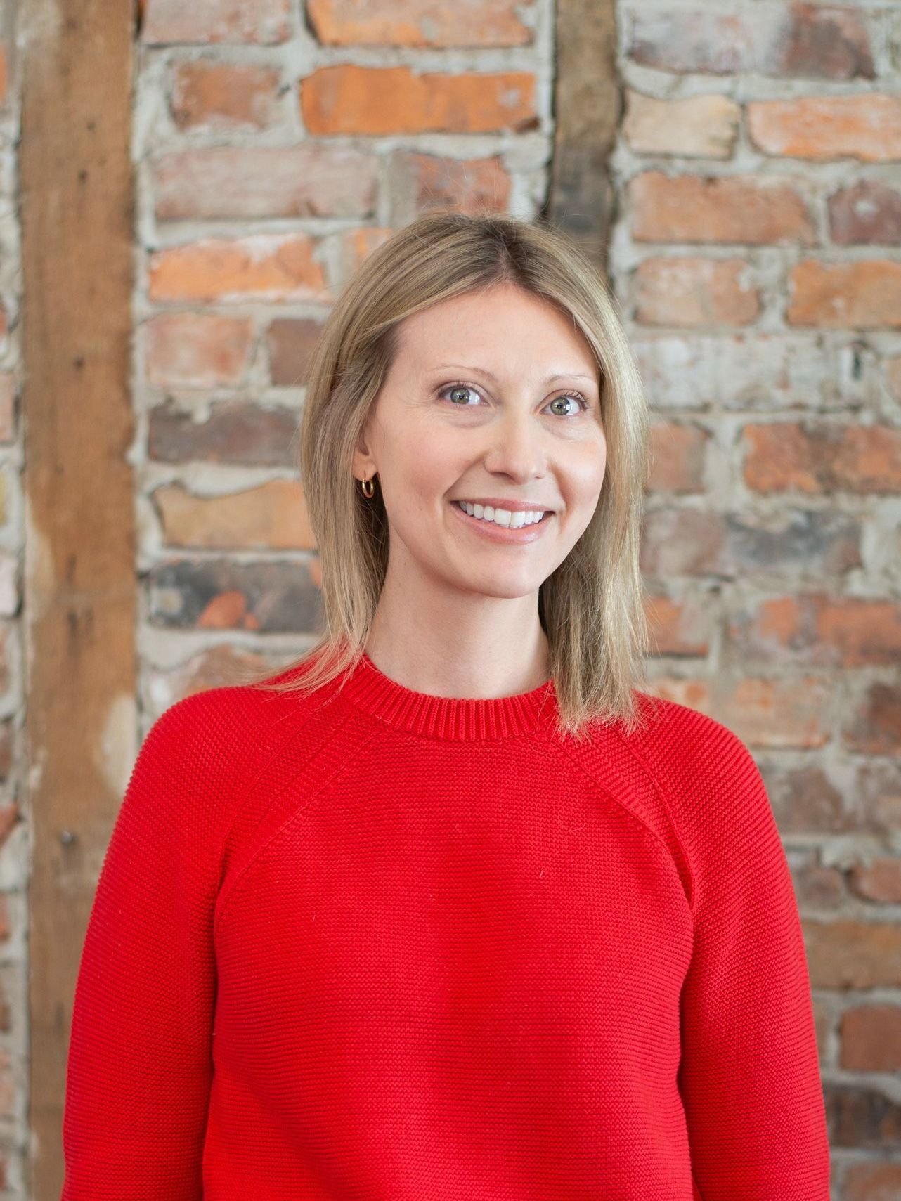 A woman in a red sweater is smiling in front of a brick wall.