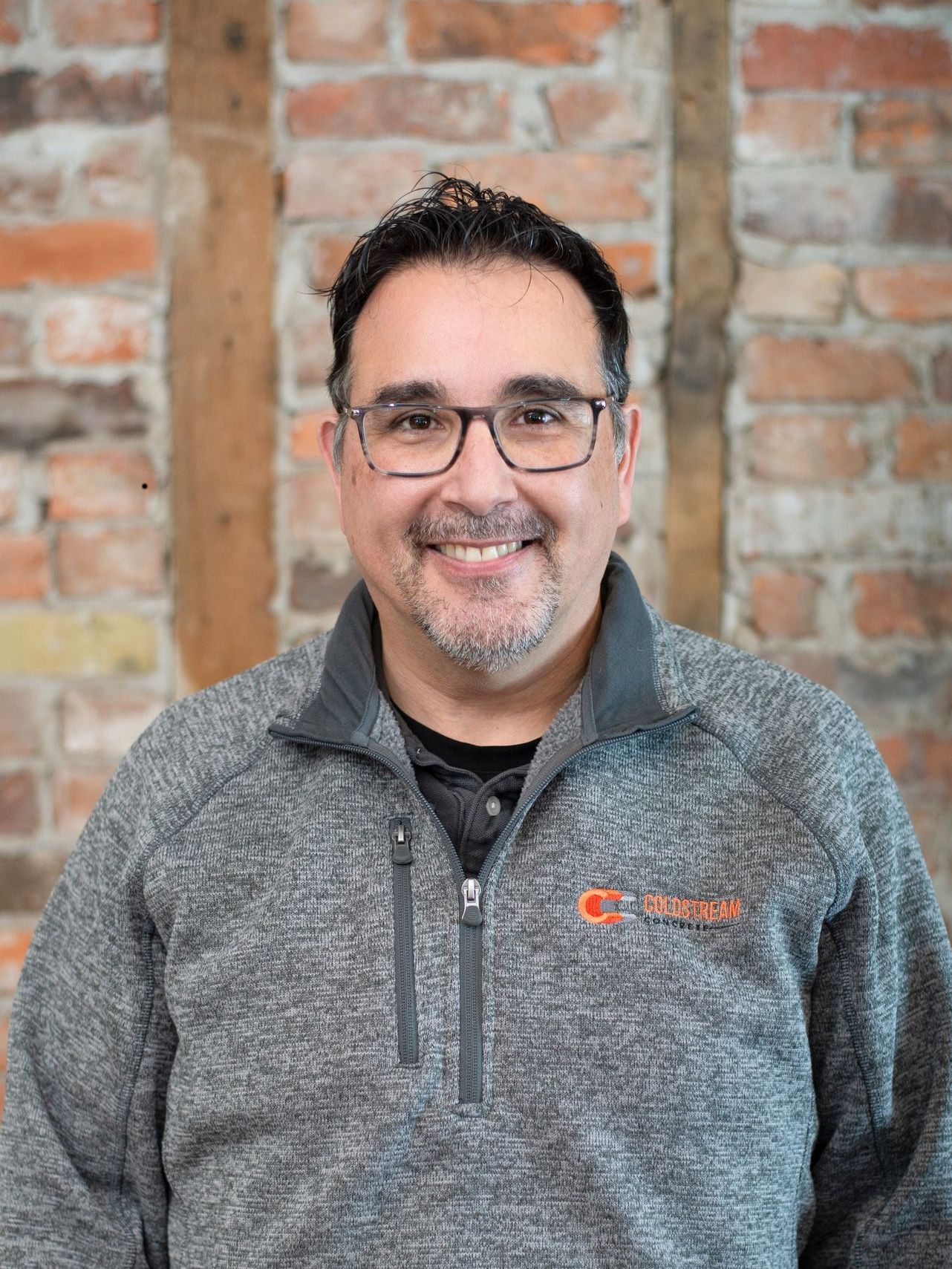 A man wearing glasses and a gray jacket is smiling in front of a brick wall.