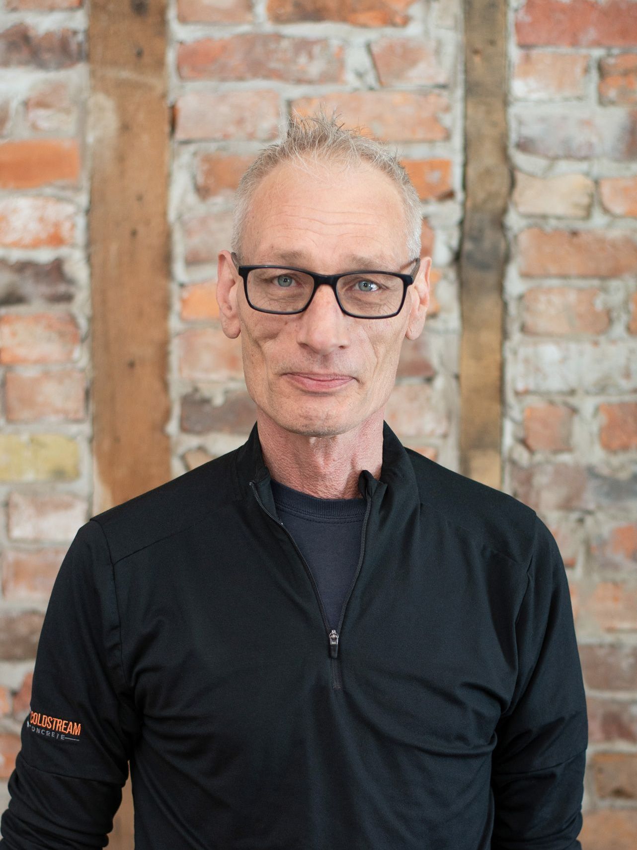 A man wearing glasses and a black shirt is standing in front of a brick wall.