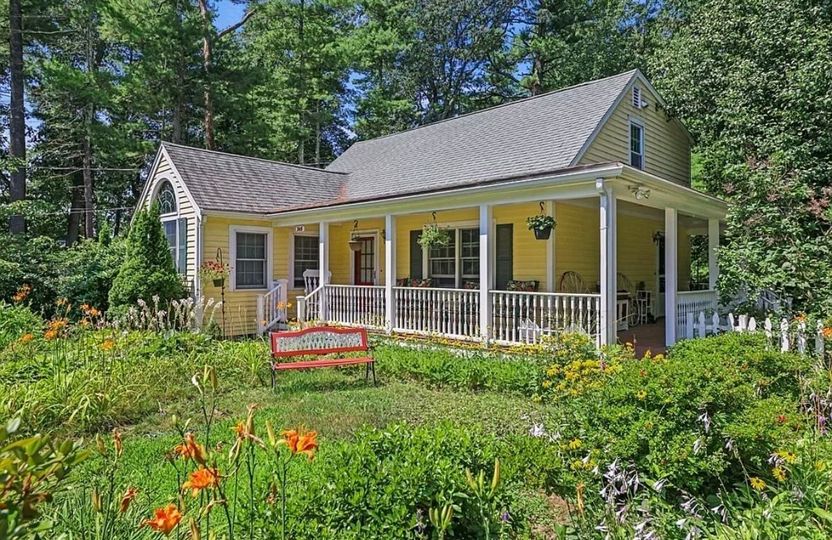 A yellow house with a porch is surrounded by trees and flowers.