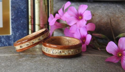 A set of two wooden rings with small purple flowers in the background. The rings are sitting on a rock. There is the corner of a small framed picture to the left.  The wooden rings are made of Cocobolo wood and they each have a center inlay of a beloved dog's ashes.