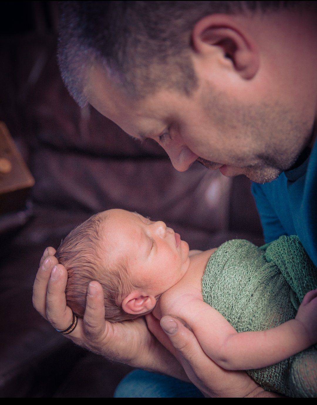 A man holding a wee babe with both hands and looking at the baby with so much love. The dad is wearing the Touch Wood Ring that holds their first child's ashes.