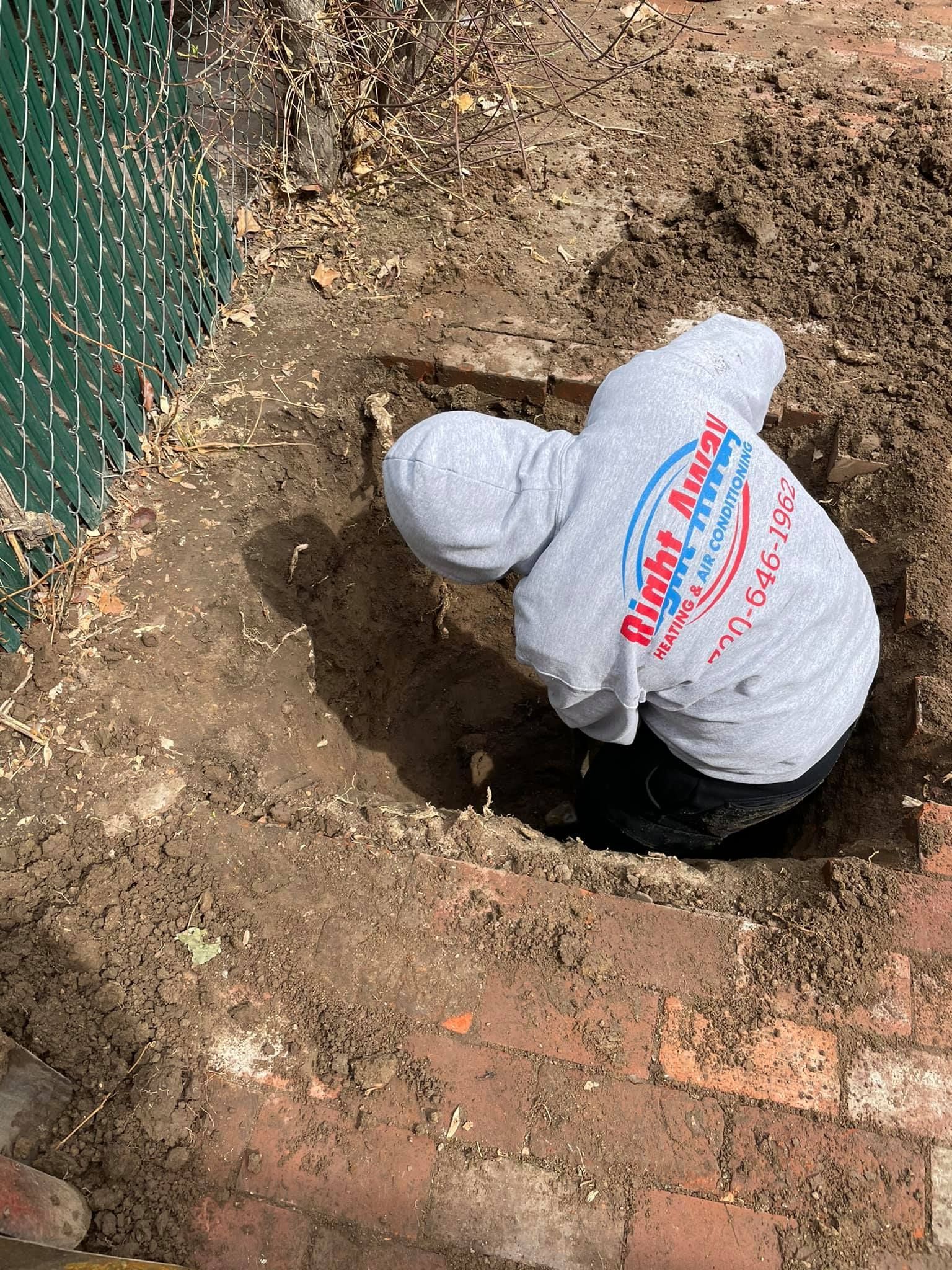 Person digging in a hole near a brick wall, wearing a gray hoodie with