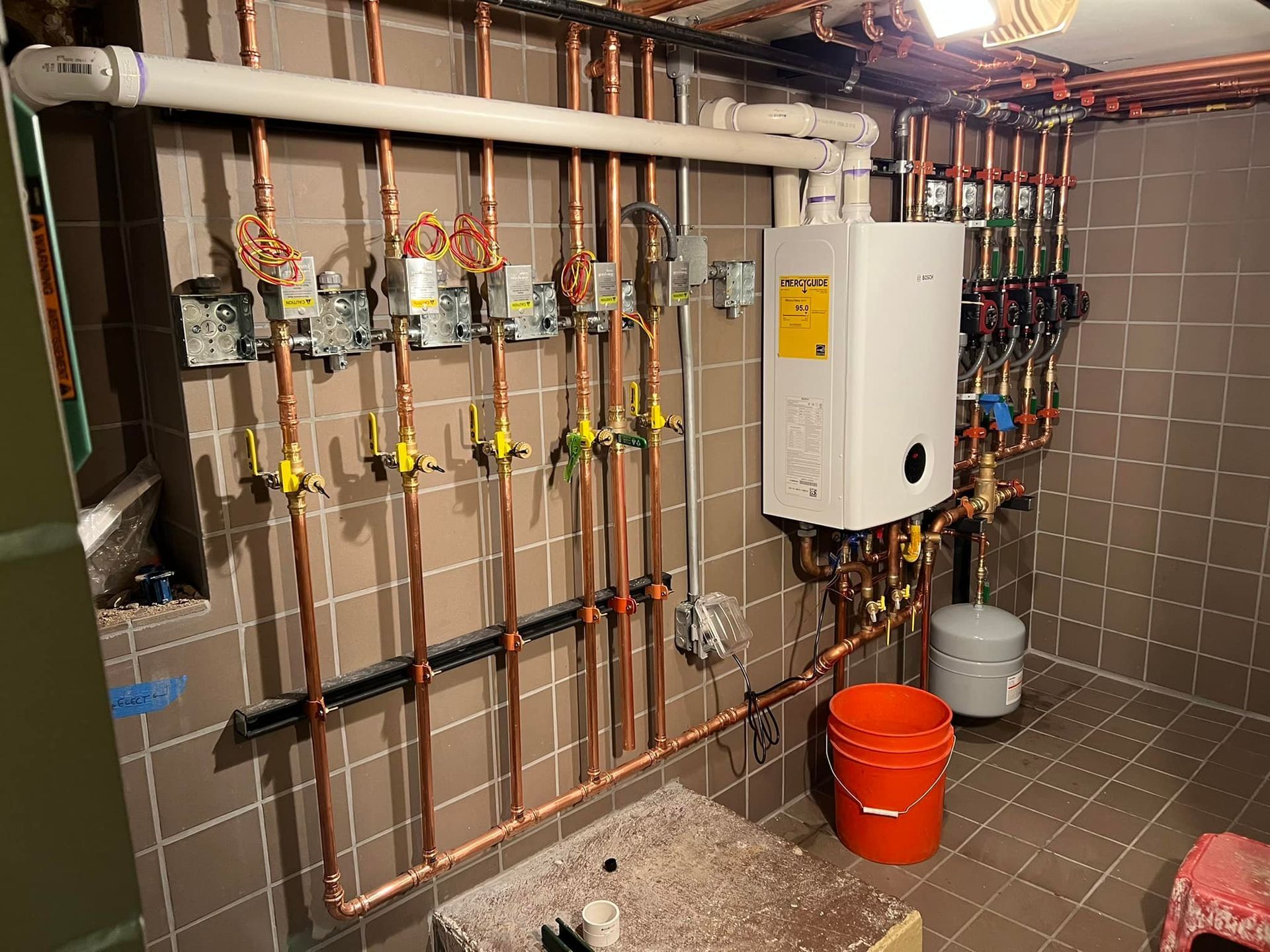 Copper pipes and heating system components mounted on a tiled wall in a basement.