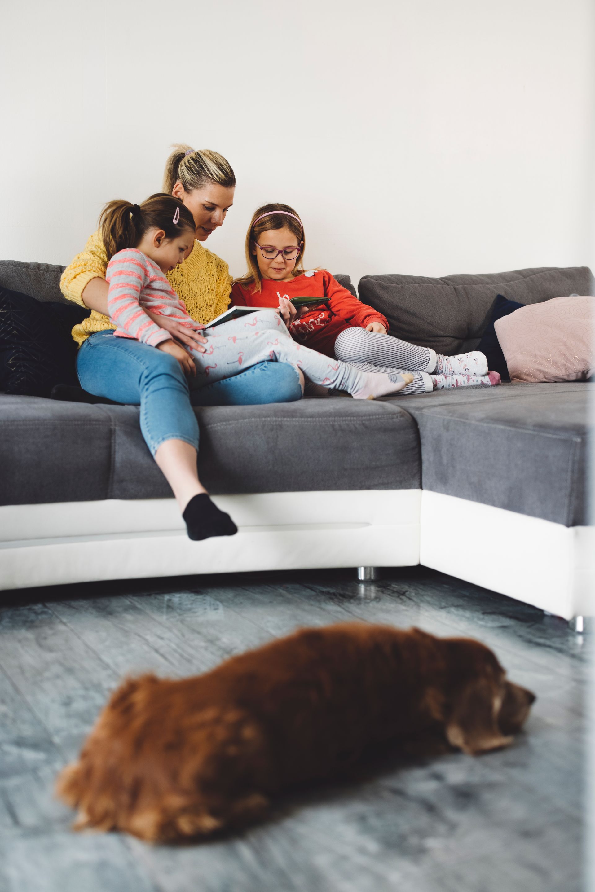 Woman and two children on a couch reading. Dog lying on the floor in front.