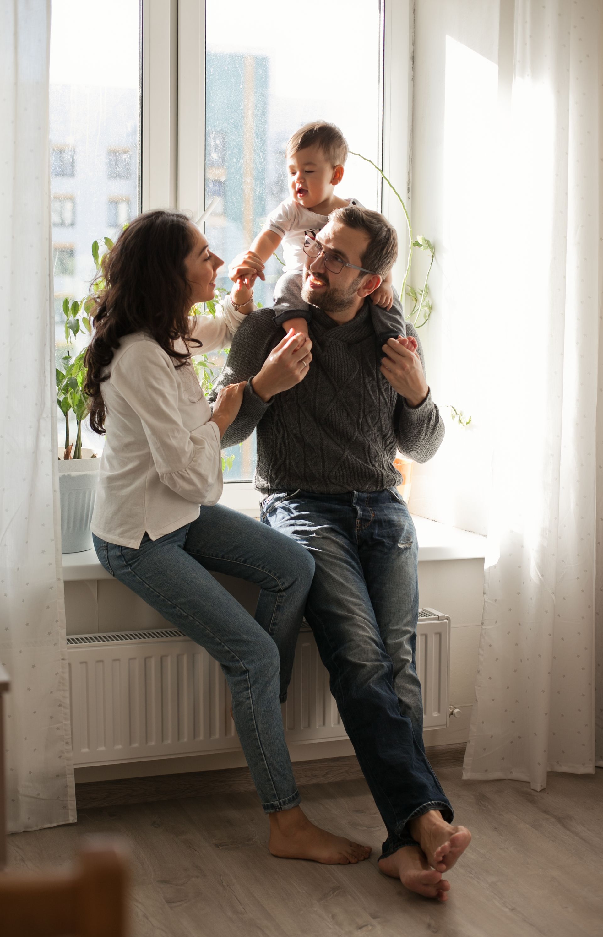 Family of three in a sunny window: mother sitting, father holding child on shoulders, all smiling.