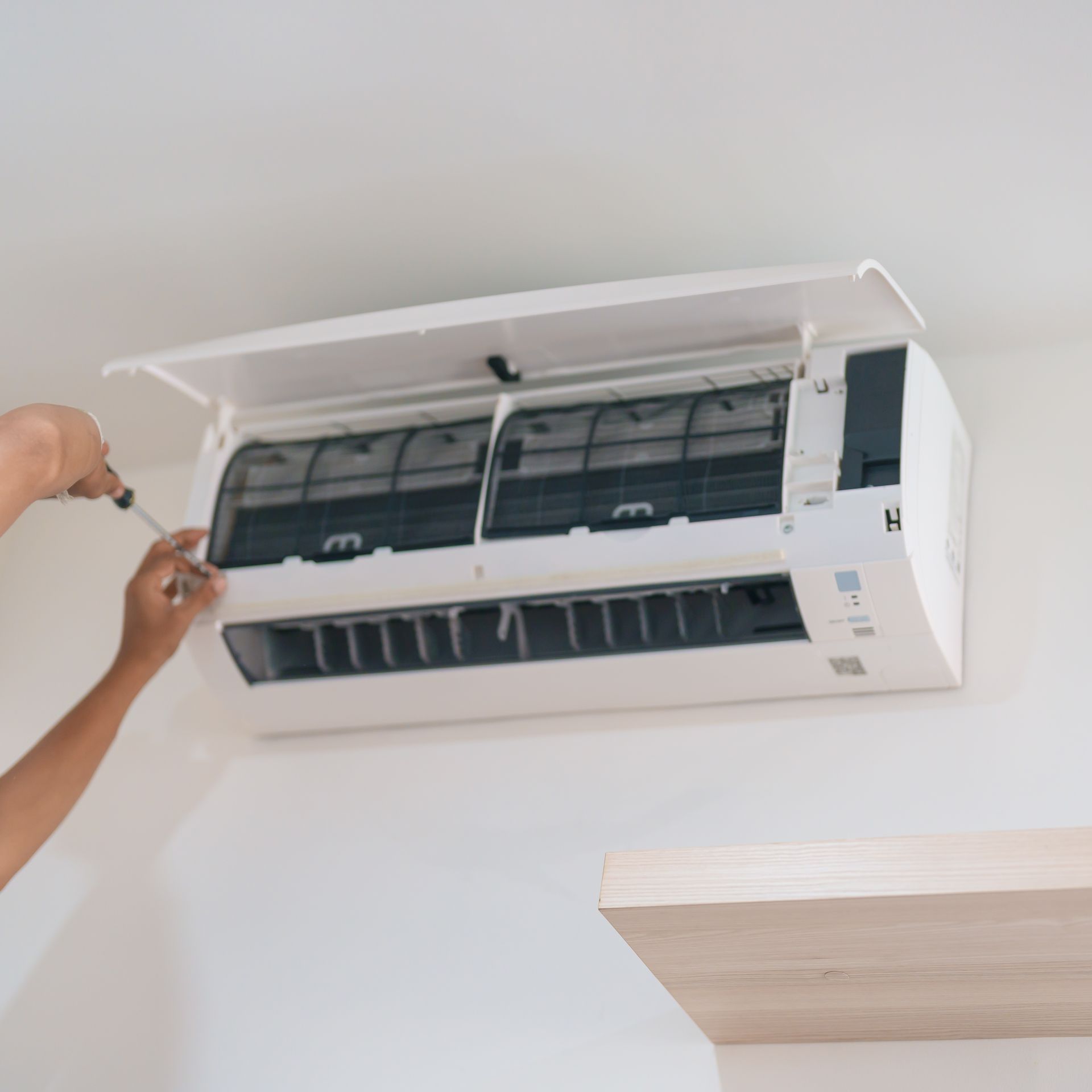 Person using a screwdriver to open a white air conditioning unit on a white wall.