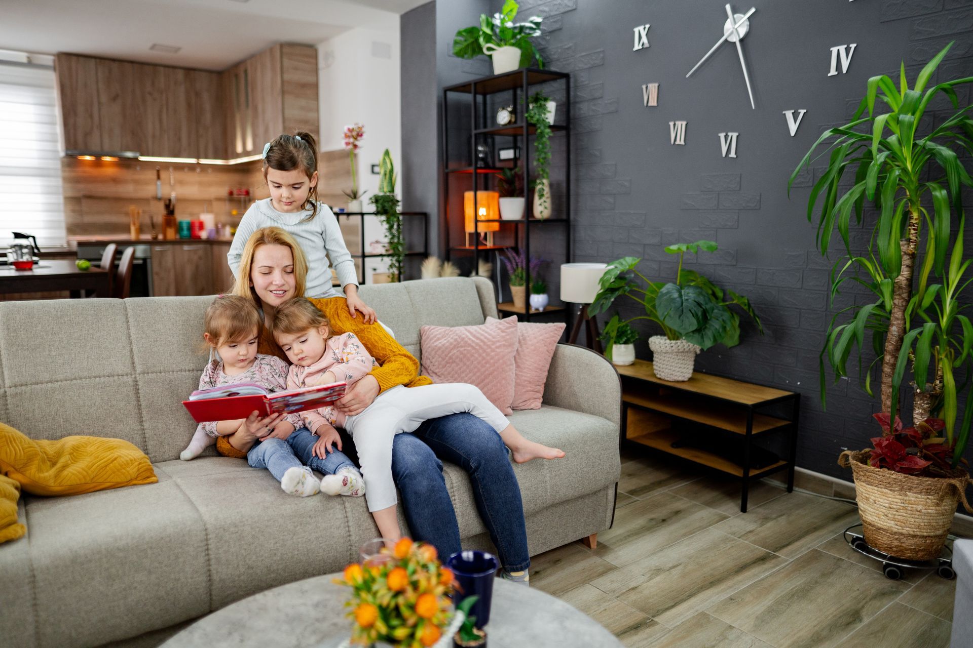 A woman and three children read on a sofa in a living room; a large clock is on a dark wall.