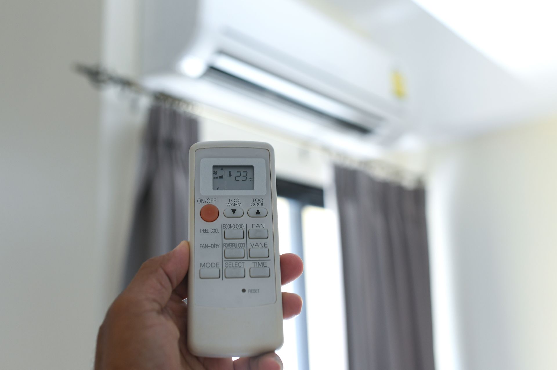 Hand holding an air conditioner remote, with the unit mounted on a white wall above a window with gray curtains.