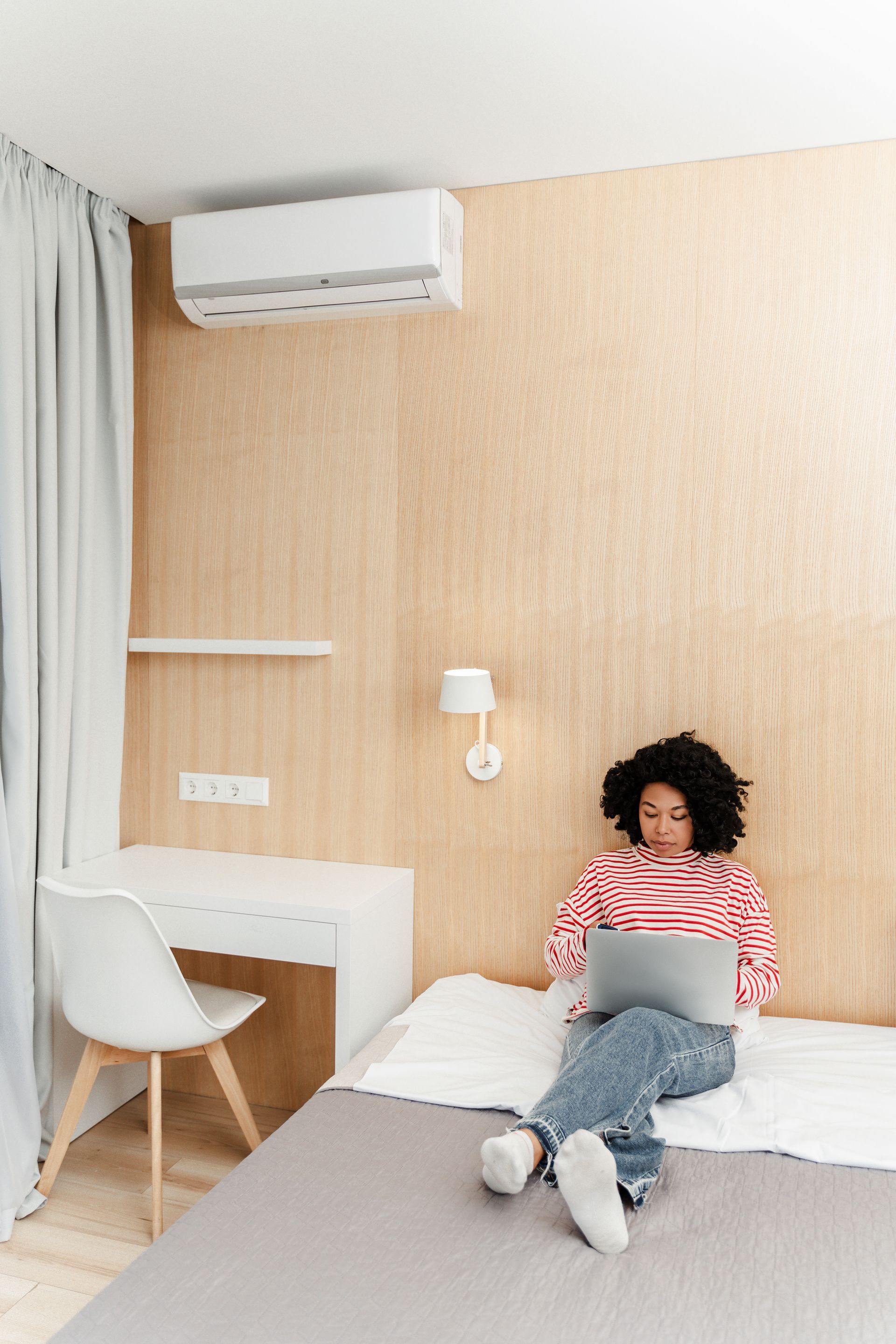 Person sits on bed working on laptop in a small room with light-colored walls, desk, and air conditioner.